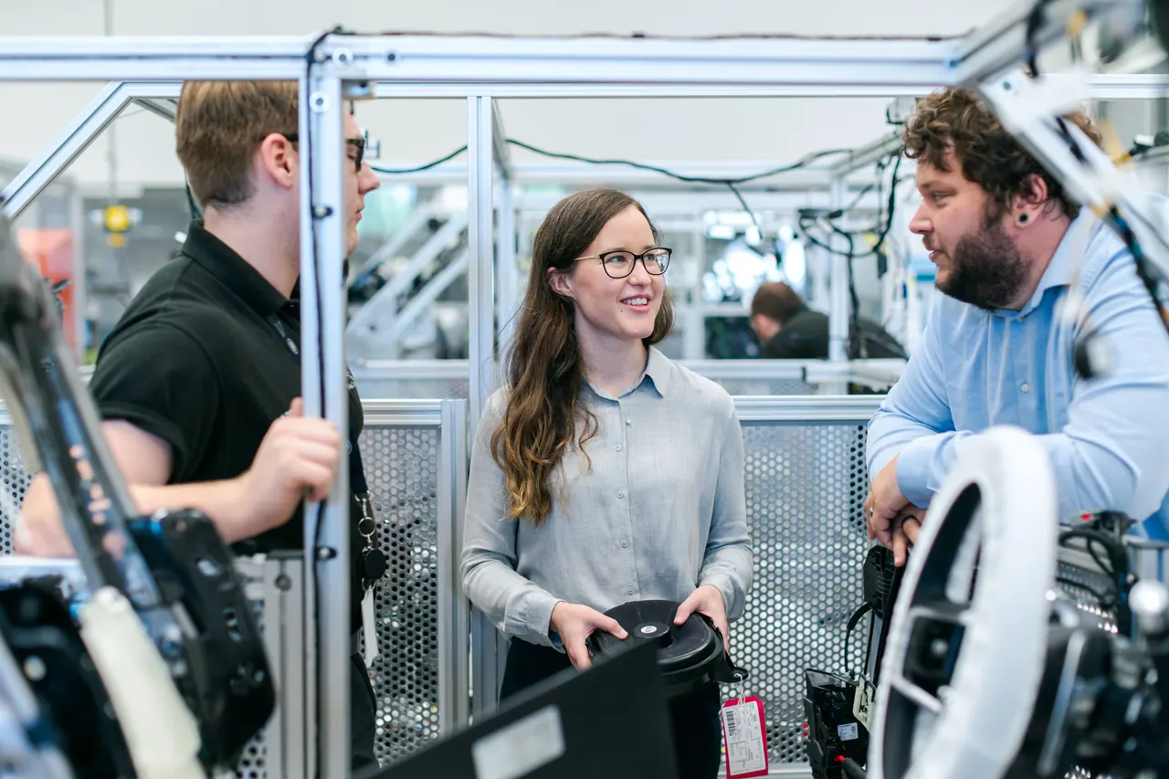 Drei Menschen besprechen ein Bauteil in einer technischen Werkstatt.